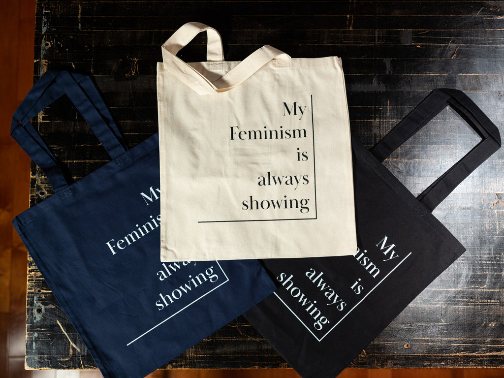 Three cotton tote bags displayed flat on black table top. Top one is a light cream colour, bottom left is navy blue, and bottom right is black. All three have the same wording on the front: My Feminism is always showing.