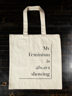 Natural cotton tote bag displayed flat on black table top. Light cream colour with black text that reads: My feminism is always showing. Each word is aligned to the right, with a black line to the right and under the text. 